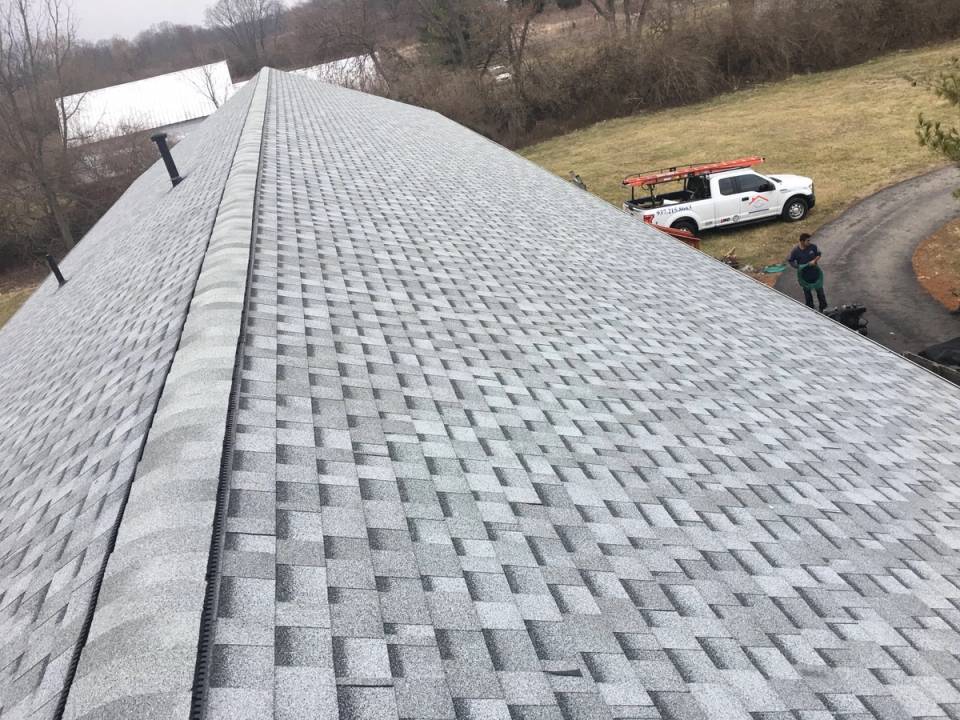 Finished asphalt shingles running the full length of a large roof, seen down the capped ridge, with a roofing truck parked on the drive below.